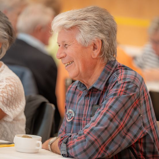 Ein älterer Mann in einem karierten Hemd sitzt am Tisch, lächelt und ist möglicherweise in ein Gespräch vertieft. Hinter ihm ist eine weitere Person verschwommen, und der Hintergrund umfasst einen Tisch und Stühle.