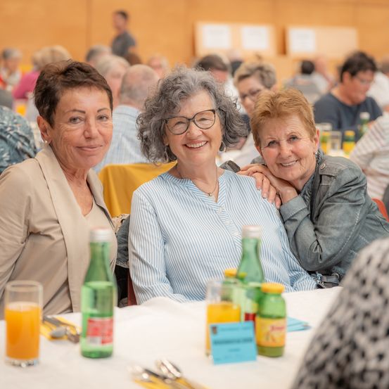 Drei ältere Frauen lächeln und posieren für ein Foto bei einem Treffen, umgeben von einem Tisch mit Getränken und einem verblassten Hintergrund mit anderen Leuten.