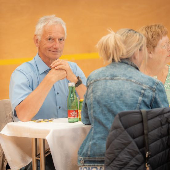 Ein älterer Mann in einem blauen Hemd sitzt an einem Tisch mit einer grünen Flasche. Er lächelt und hat die Hände auf dem Kinn. Hinter ihm schaut eine Frau mit blonden Haaren zu ihm. Eine weitere Frau mit Brille sitzt hinter ihr.