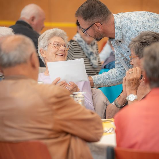 Eine ältere Frau lächelt, während sie ein Blatt Papier in der Hand hält, während sich ein Mann mit einem Stück Papier über sie beugt. Der Raum ist mit anderen älteren Menschen gefüllt, die alle an einem Tisch sitzen.