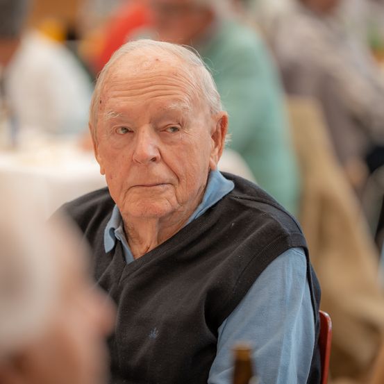Ein älterer Mann sitzt an einem Tisch und trägt ein blaues Hemd und eine schwarze Weste. Er hat einen ernsten Gesichtsausdruck, möglicherweise in einer Unterhaltung vertieft. Hinter ihm sitzen weitere Personen, verschwommen und unscharf.