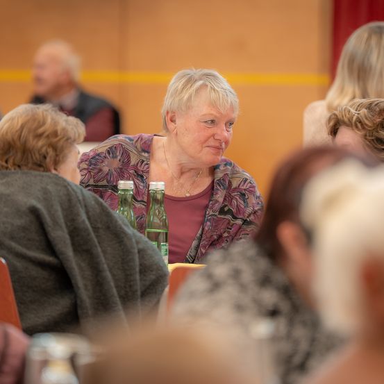 Eine ältere Frau mit blondem Haar sitzt mit zwei grünen Flaschen am Tisch, während andere um sie herum sitzen. Eine Person mit grauem Haar ist in der Nähe.