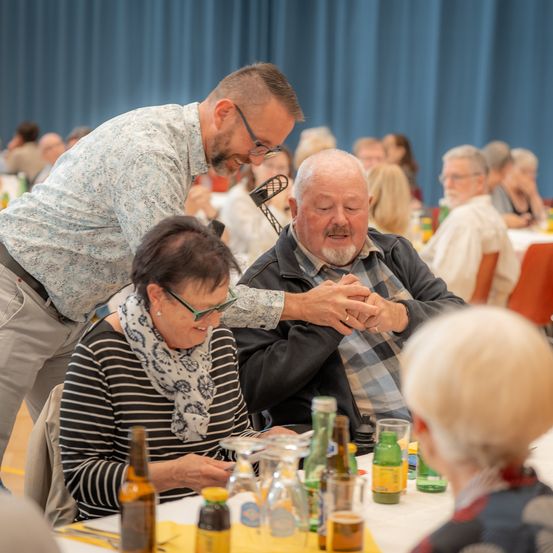 Ein älterer Mann lächelt, während eine ältere Frau mit einem Schal neben ihm an einem Tisch mit verschiedenen Getränken sitzt. Ein Mann mit Brille beugt sich über sie.