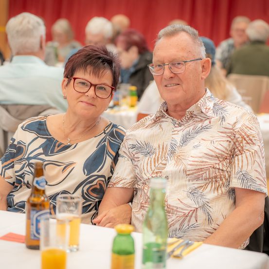 Ein älteres Paar mit Brille sitzt an einem Tisch in einem Restaurant. Flaschen und Gläser stehen auf dem Tisch, und andere Menschen sind im Hintergrund sitzend.