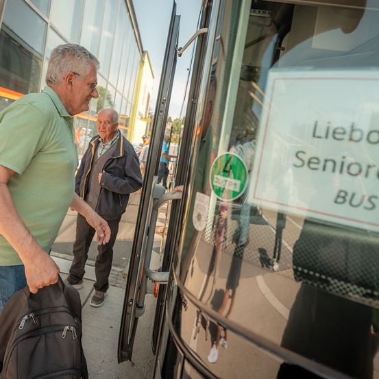 Zwei ältere Männer steigen in einen Bus ein, der mit 'Liebo Senior BUS' beschriftet ist. Einer hält einen Rucksack und der andere lächelt. Im Hintergrund befindet sich ein Gebäude mit Glasfenstern.