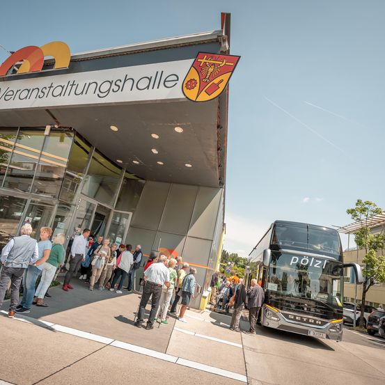 Ein Doppeldeckerbus steht vor einem modernen Veranstaltungsgebäude. Viele Menschen, einige mit Taschen, versammeln sich um den Eingang des Busses.