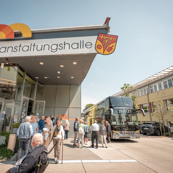 Mehrere Menschen stehen vor einem Gebäude mit der Aufschrift 'Anstaltungshalle', vor dem ein Bus geparkt ist. Der Bus trägt den Schriftzug 'Poliz' auf der Vorderseite. Eine Person mit Rucksack sitzt auf dem Boden.