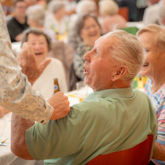 Ein älterer Mann in einem grünen T-Shirt lacht herzlich, während er von einer anderen Person spielerisch geschlagen wird, umgeben von einer lebhaften Menge.