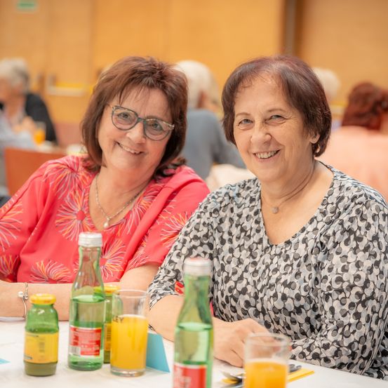 Zwei Frauen, die lächeln und an einem Tisch mit Getränken und Essen sitzen, möglicherweise bei einem gesellschaftlichen Ereignis. Sie tragen Brillen und scheinen für ein Foto zu posieren.