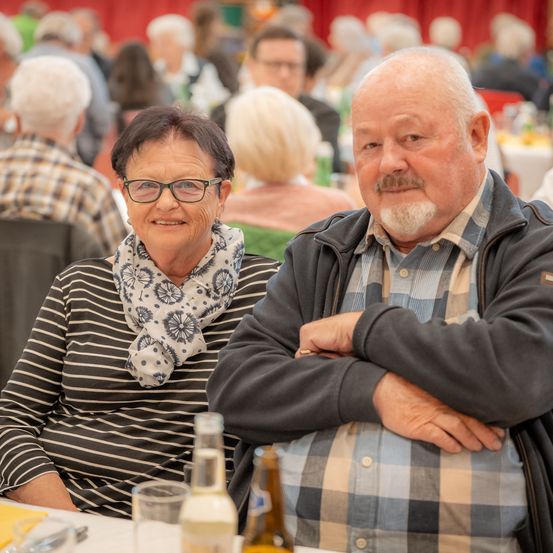 Zwei ältere Personen, ein Mann und eine Frau, sitzen zusammen an einem Tisch mit Getränken. Sie lächeln und scheinen eine gesellige Zusammenkunft zu genießen. Der Mann hat die Arme verschränkt, während die Frau eine Schürze trägt. Sie sind von anderen Personen umgeben, die an Tischen sitzen.