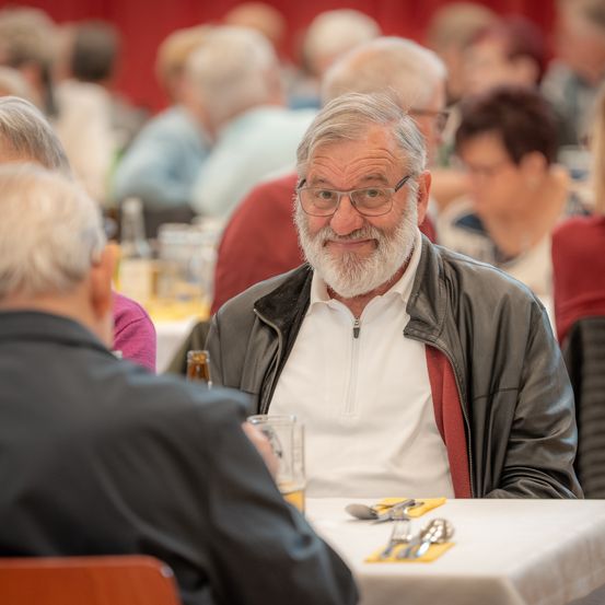 Ein älterer Mann mit Bart und Brille sitzt an einem Tisch und trägt eine Lederjacke. Er scheint sich mit einem anderen Mann zu unterhalten. Der Tisch ist mit einem Glas, Besteck und einer Serviette gedeckt. Hinter ihm sitzen weitere Personen, die in ihre eigenen Gespräche vertieft sind.