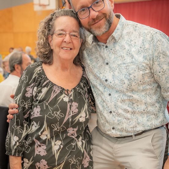 Ein lächelndes Paar steht in einem Raum zusammen, mit einem unscharfen Hintergrund anderer Leute. Die Frau trägt eine Bluse mit Blumenmuster und eine Brille, während der Mann ein gemustertes Hemd und eine Brille trägt. Sie scheinen für ein Foto zu posieren.