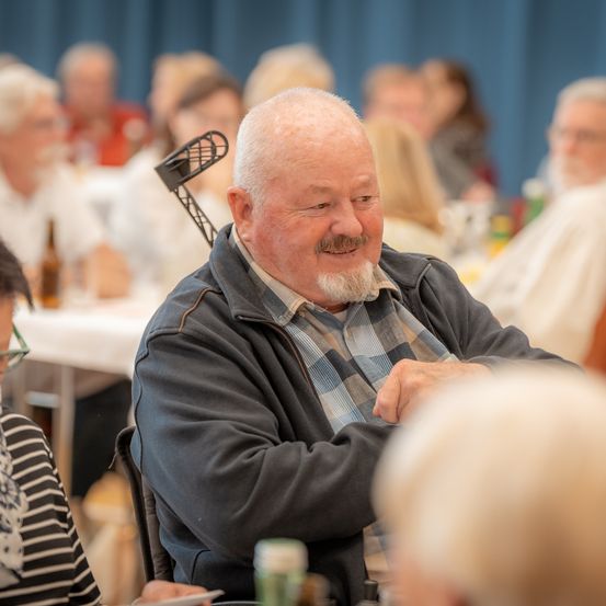 Ein älterer Mann mit weißem Bart und Schnurrbart sitzt lächelnd an einem Tisch mit anderen Leuten.