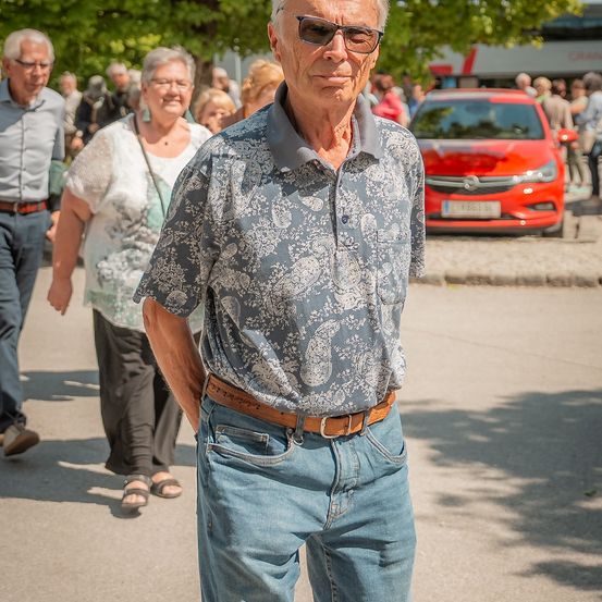 Ein älterer Mann in einem gemusterten Hemd und Jeans läuft neben zwei älteren Erwachsenen auf einer sonnigen Straße mit einem roten Auto und Menschen im Hintergrund.