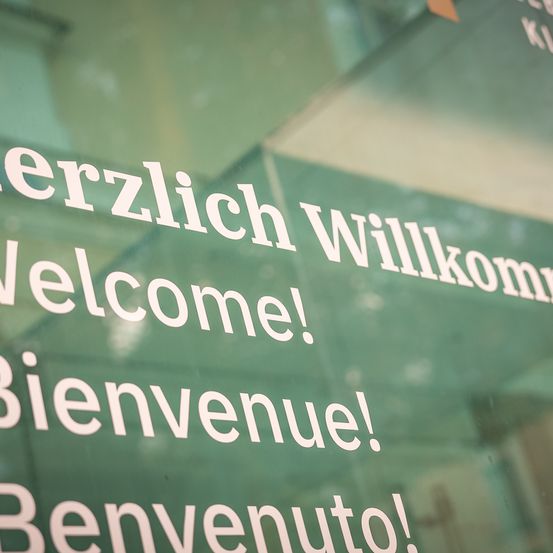 Eine Nahaufnahme einer Glastür zeigt mehrsprachige Willkommensschilder. Von oben nach unten steht Herzlich Willkommen, Welcome!, Bienvenue! und Benvenuto!