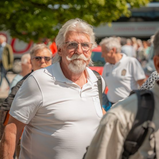 Ein älterer Mann mit weißem Haar und einem Schnurrbart trägt eine Brille und ein weißes Poloshirt. Er läuft in einer Menschenmenge. Hinter ihm sind andere Menschen zu sehen, und im Hintergrund ist teilweise ein Bus sichtbar.