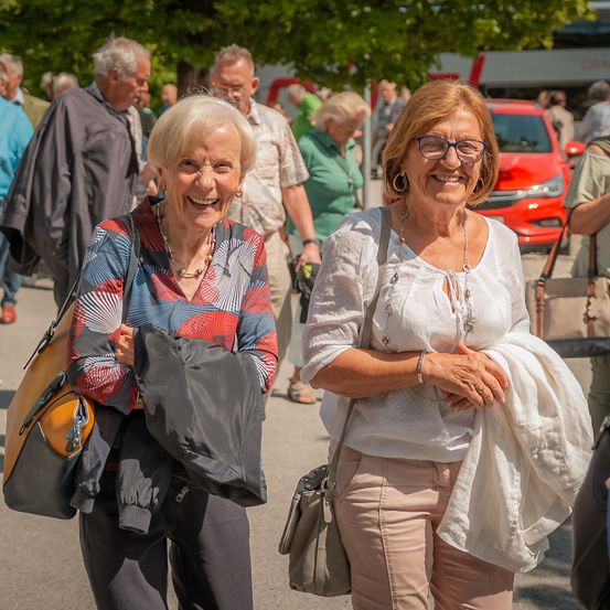 Zwei ältere Frauen lächeln und gehen in einem geschäftigen Außenbereich. Eine trägt ein farbenfrohes Oberteil und eine schwarze Jacke. Hinter ihnen gehen mehrere Menschen, und ein rotes Auto ist geparkt. Bäume und ein Bus sind im Hintergrund.