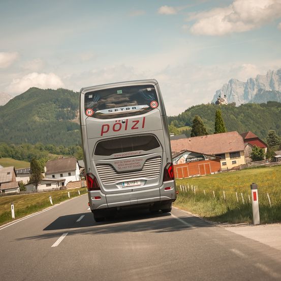 Ein silberner Bus mit dem Wort 'Poizl' auf der Rückseite fährt auf einer Straße mit Bergen im Hintergrund.