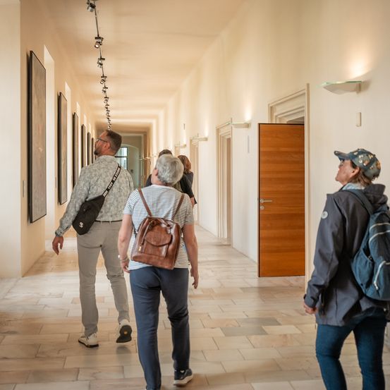 Eine Gruppe von vier Personen geht einen Flur in einem Gebäude entlang. Drei von ihnen tragen Rucksäcke.