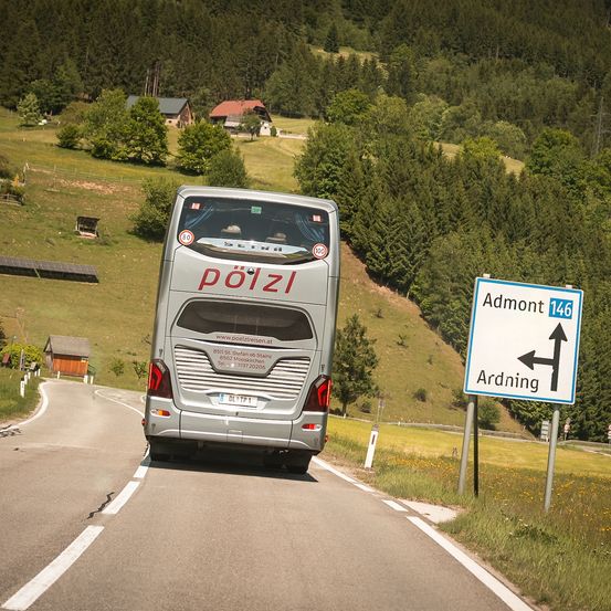 Ein Bus namens Pölzli fährt auf einer Straße, an der ein Schild auf Admont und Ardning hinweist. Im Hintergrund sind Bäume und Häuser zu sehen.