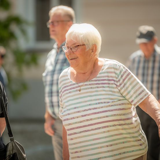 Eine ältere Frau mit gestreifter Bluse und Brille geht mit zwei Männern im Freien, einer trägt einen Hut. Sie scheinen sich in einem Innenhof zu befinden.