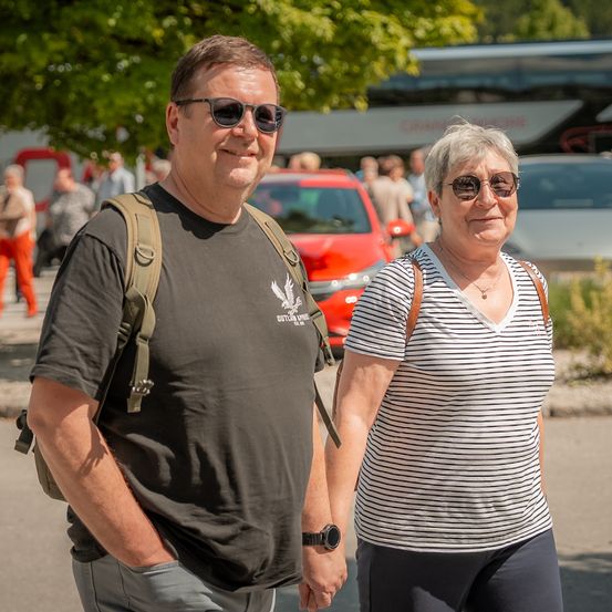 Ein älteres Paar geht lächelnd die Straße entlang, mit einem roten Auto und einem Bus im Hintergrund.