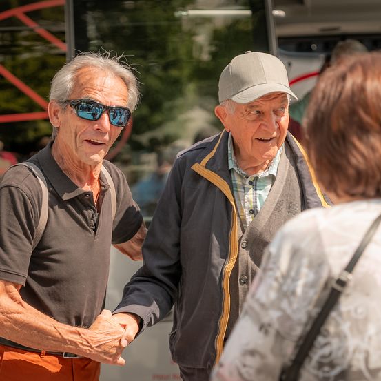 Zwei ältere Männer schütteln sich draußen die Hände. Einer trägt eine Sonnenbrille und einen Rucksack, der andere eine Mütze und eine Jacke. Eine Frau steht in der Nähe.