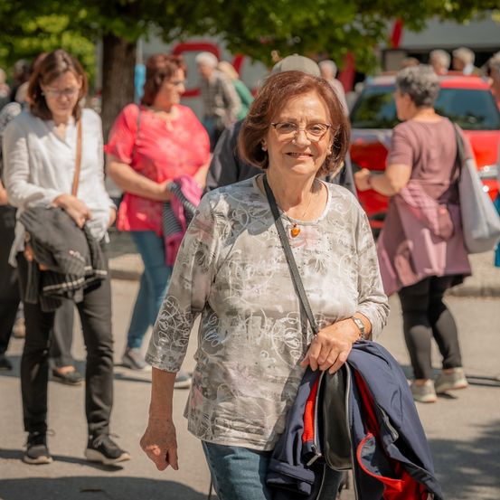 Eine ältere Frau mit Brille lächelt und geht die Straße entlang, eine Jacke tragend, mit mehreren Personen und einem Auto im Hintergrund.