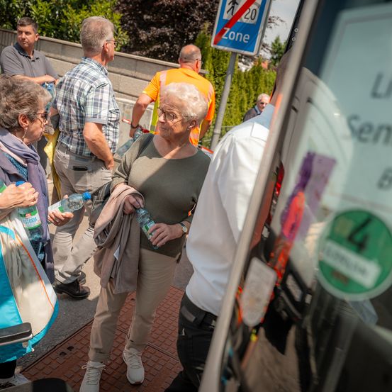 Eine Gruppe älterer Menschen steht in einer Schlange. Eine von ihnen hält zwei Flaschen Wasser. Dahinter weist ein Schild auf ein 20 km/h Tempolimit und eine Sperrzone hin.