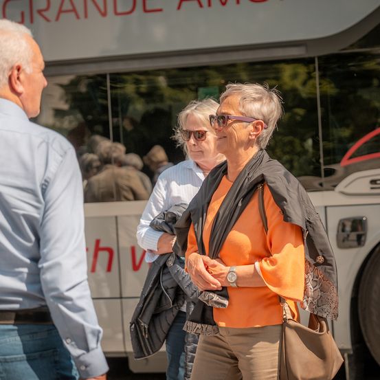 Eine Gruppe von Menschen steht draußen in der Nähe eines Busses, eine Frau trägt ein orangefarbenes Shirt und eine andere hält eine schwarze Jacke.
