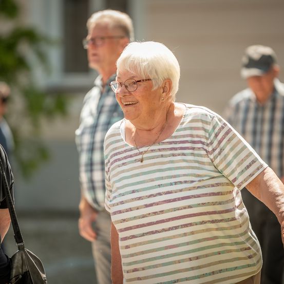 Eine ältere Frau mit Brille und einer Halskette lächelt und geht draußen mit zwei Männern in karierten Hemden.