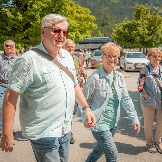 Eine Gruppe älterer Menschen, darunter ein lächelnder Mann und eine Frau, die Händchen halten, gehen an einem sonnigen Tag im Freien. Hinter ihnen sind mehrere Autos geparkt, und es gibt Bäume und ein Gebäude im Hintergrund.