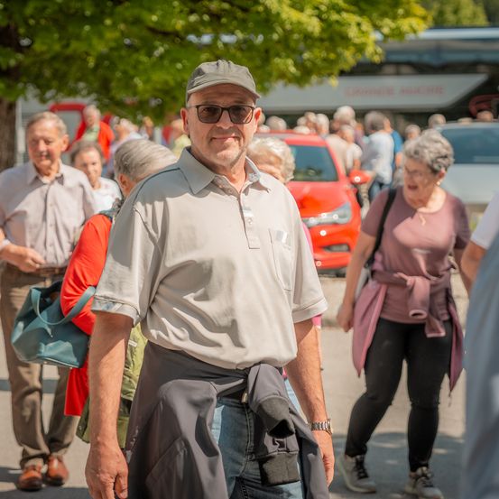Ein Mann mit einer Kappe und Sonnenbrille steht auf einer belebten Straße, um ihn herum gehen Autos und Menschen. Im Hintergrund befinden sich Bäume und geparkte Fahrzeuge.