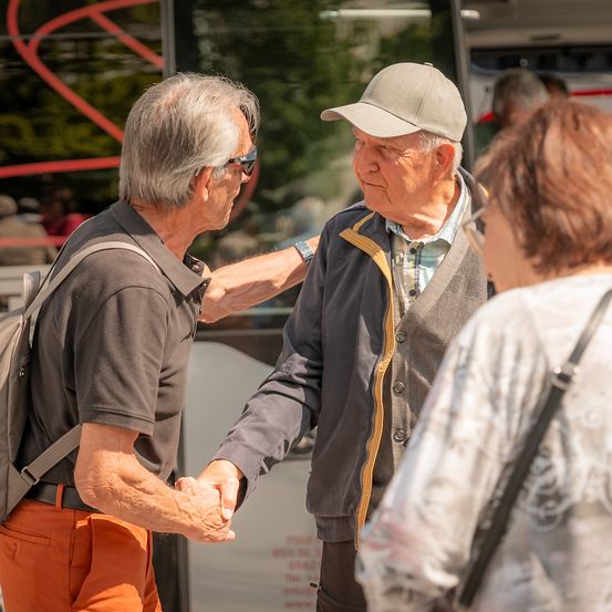 Zwei ältere Männer, einer mit einer Mütze, schütteln sich die Hände, während sie neben einem Bus stehen. Eine ältere Frau hört ihnen zu. Im Hintergrund ist ein Bus mit einem roten Design zu sehen.