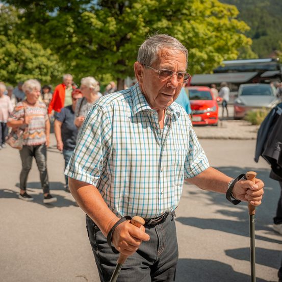 Ein älterer Mann geht draußen mit zwei Stöcken, trägt ein kariertes Hemd. Im Hintergrund gehen mehrere Menschen und es sind Autos geparkt. Bäume und Berge sind sichtbar.
