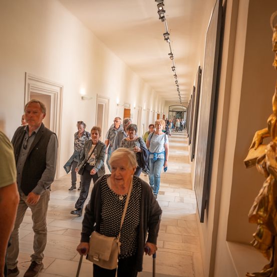 Eine Gruppe von Menschen geht in einem langen Flur. Einige von ihnen sind älter, und einige tragen Taschen. Es gibt Bilderrahmen an der Wand, und eine Statue befindet sich auf der rechten Seite.