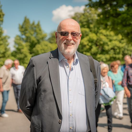 Ein älterer Mann mit weißem Bart und Sonnenbrille lächelt in einem Park, wo andere spazieren gehen.