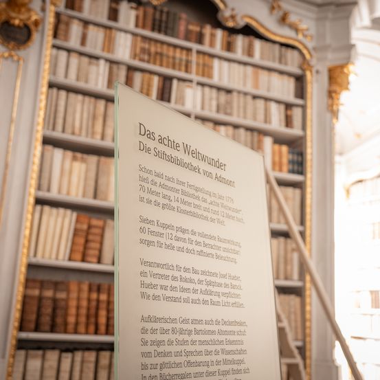 Ein Schild vor einer Bibliothek zeigt Informationen über ihre Architektur. Die Bibliothek ist mit vielen Büchern auf Regalen gefüllt. Das Schild informiert über die architektonischen Wunder der Bibliothek.