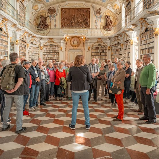 Eine Gruppe von Menschen steht in einer Bibliothek mit verzierten Decken und Wänden und hört einer Frau in der Mitte zu. Die Bibliothek hat einen geometrischen Boden und ist mit Bücherregalen gefüllt.