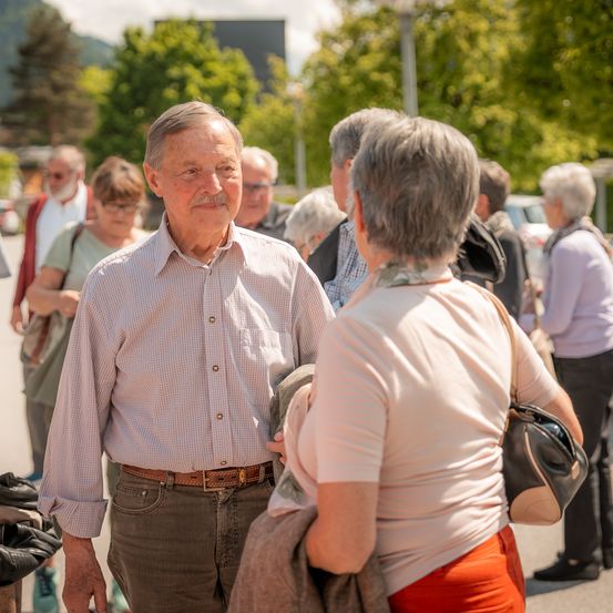 Eine Gruppe älterer Menschen steht draußen, vielleicht im Gespräch. Sie tragen alle Freizeitkleidung. Der Mann in der Mitte hat einen Schnurrbart und trägt einen Gürtel. Die Frau rechts trägt eine Handtasche. Im Hintergrund befinden sich Bäume und ein Gebäude.