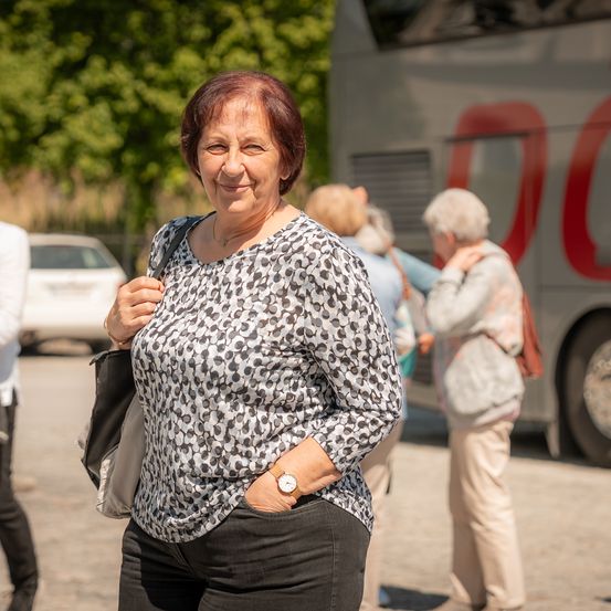 Eine ältere Frau mit einem schwarz-weißen Shirt lächelt in die Kamera, während sie vor einem Bus geht. Andere Leute laufen hinter ihr.