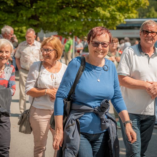 Eine Gruppe lächelnder Erwachsener in Freizeitkleidung geht auf einer Stadtstraße. Eine Frau in Blau führt, trägt eine Halskette und eine Tasche. Dahinter folgen eine weitere Frau in Weiß und ein Mann in Weiß und mit einer Brille. Im Hintergrund befinden sich Bäume und ein Bus.