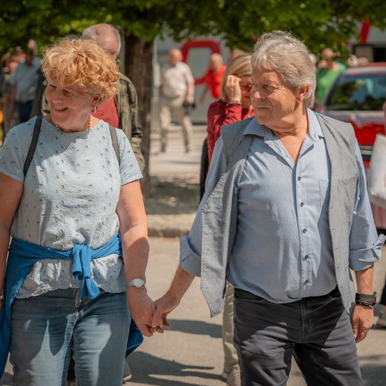 Ein älteres Paar geht Hand in Hand durch eine sonnige Außenumgebung, mit anderen Menschen und Autos im Hintergrund.