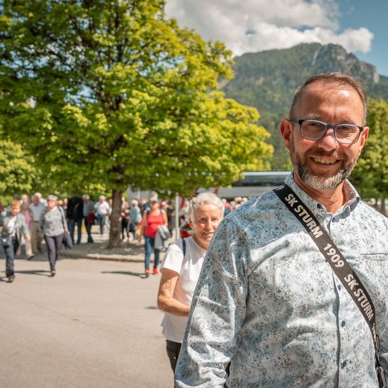 Ein lächelnder Mann mit Brille steht in einem Park mit Bergen im Hintergrund, während andere vorbeigehen.