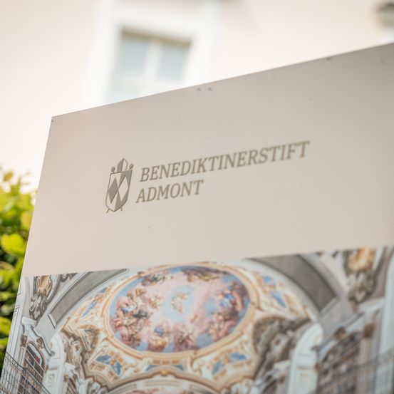 Ein weißes Schild zeigt das Logo und den Namen 'Benediktinerstift Admont' vor dem Hintergrund eines historischen Gebäudes und einer reich verzierten Decke.
