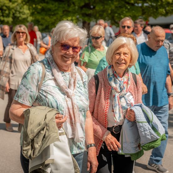 Mehrere Menschen, darunter zwei ältere Frauen, gehen in einer Außenumgebung. Sie tragen Sonnenbrillen und haben Jacken bei sich. Im Hintergrund sind Bäume und eine Menschenmenge zu sehen.