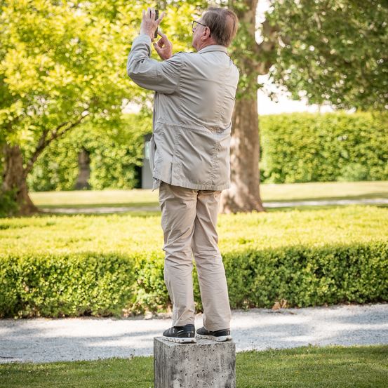 Ein älterer Mann macht ein Foto in einem Park, steht auf einem Betonblock. Er trägt eine leichte Jacke und Khakihosen. Hinter ihm befinden sich Hecken und Bäume.