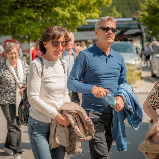 Ein Paar geht in der Stadt, die Frau hält eine Jacke und der Mann eine Wasserflasche. Sie tragen beide Sonnenbrillen. Hinter ihnen sind andere Leute und Fahrzeuge.