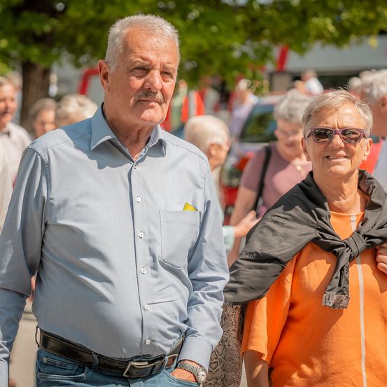 Ein älterer Mann und eine ältere Frau stehen lächelnd draußen. Der Mann trägt ein blaues Hemd und einen schwarzen Gürtel. Die Frau hat eine Halskette und eine Sonnenbrille. Unscharfe Menschen und Autos sind im Hintergrund.