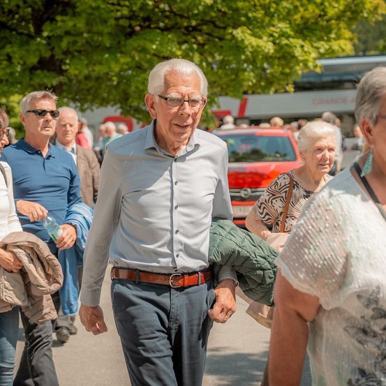 Ein älterer Mann mit Brille geht mit anderen auf einer sonnigen Straße. In der Nähe trägt eine Frau eine grüne Jacke. Im Hintergrund sind ein rotes Auto und ein Bus zu sehen.